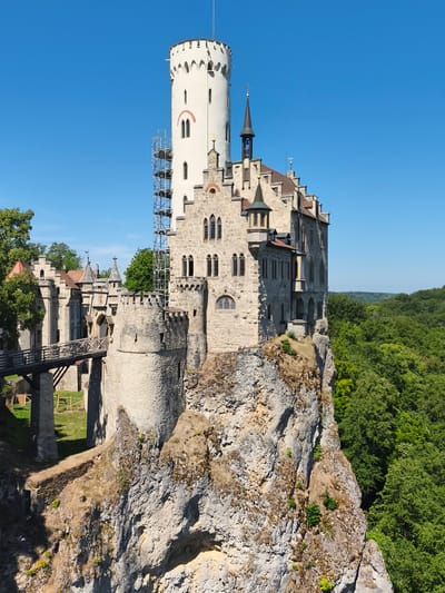 The image shows Lichtenstein Castle, a neo-Gothic castle located in the Honau, Germany. The castle is perched atop a sheer rock face, surrounded by lush green forest. The main structure features a tall, white tower with arched windows and battlements, constructed from stone blocks. Adjacent to it is a lower section of the castle, also built of stone, with smaller towers and a steep, tiled roof. A wooden bridge extends from the lower castle, connecting to a round stone tower to the left. Scaffolding is visible on the side of the white tower, suggesting ongoing maintenance or construction. The sky is a clear, bright blue, indicating a sunny day. The overall scene depicts a historic and picturesque castle in a dramatic natural setting.