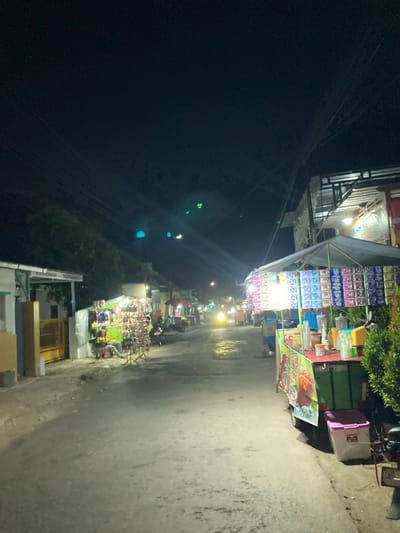 The image depicts a nighttime street scene in Proyonangg, Indonesia, with several stalls set up along the roadside. The street is paved and appears wet, reflecting the ambient light. On the left side of the street, a building with a white facade and a yellow gate is visible, with racks of merchandise displayed outside. Further down the street, more stalls are illuminated, featuring various products, including packaged goods and what appear to be food or drink items. A person is seated near one of the stalls. On the right side of the street, a larger stall with a canopy is prominent, displaying rows of colorful packages. Several motorbikes are parked in the vicinity, and a person is operating a food cart. The overall atmosphere suggests a lively market or street food area. The sky above is dark, with visible power lines.