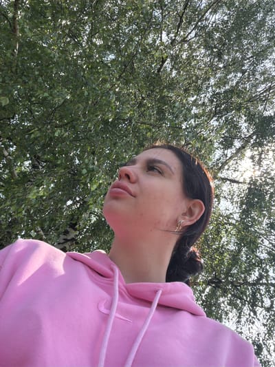 A medium shot shows a young woman wearing a pink hoodie, looking up through a canopy of green leaves. The camera angle is from below, looking upwards towards the sky. The woman's face is visible in profile, with her eyes looking upwards. Her hair is dark and pulled back. She has a small earring in her left ear. The leaves of the trees are dense and partially obscure the bright sky, with sunlight filtering through the branches. The background consists of trees and sky, suggesting an outdoor setting. The image does not contain any specific details indicating a location in the city of Чумна, Russia.