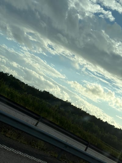The image displays a landscape scene, likely captured from a moving vehicle, as indicated by the tilted perspective and the presence of a road barrier and asphalt in the foreground. The upper portion of the image is dominated by a dramatic sky filled with a mix of light and dark clouds, suggesting dynamic weather conditions, possibly during sunrise or sunset. The clouds are layered and textured, with patches of blue sky visible between them. Below the sky, a dense line of dark green trees forms a treeline along a gently sloping hill. In front of the treeline, tall green grass and vegetation are visible. A metal guardrail runs horizontally across the lower part of the image, separating the roadway from the grassy embankment. The road surface, visible in the very bottom, is dark asphalt marked with white lines. The overall impression is of a rural or semi-rural setting, possibly along a highway, with a focus on the interplay of natural elements like the sky, trees, and vegetation under a dynamic sky. No people or specific buildings are visible, and there are no discernable actions or interactions taking place. The location in Oskarsberg, Sweden, is not visually confirmed by any landmarks or textual elements within the image itself.