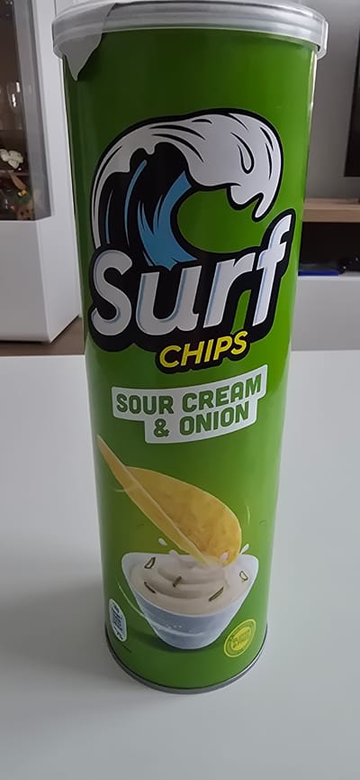 A green cylindrical canister of "Surf Chips" with "Sour Cream & Onion" flavor is displayed on a white surface. The canister features a prominent logo with a white wave cresting over blue and black water. Below the logo, the text "Surf CHIPS" is visible. Further down, "SOUR CREAM & ONION" is printed in white letters. An illustration of a potato chip being dipped into a bowl of sour cream with chives is present at the bottom of the canister, accompanied by a green circular label indicating "GLUTEN FREE". The background is blurred and shows a white wall and a glimpse of a shelf with objects. There are no people or animals present in the image. The scene appears to be indoors, possibly in a home or retail setting. The location in Haren (Ems), Germany is not discernible from the visual information provided in the image itself.