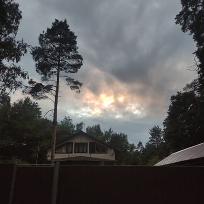 This is a medium-format, low-angle, outdoor, daylight photograph taken in Kratovo, Russia. The scene is dominated by a dark, overcast sky with patches of bright, golden-yellow light breaking through the clouds. In the foreground, a dark brown wooden fence runs horizontally across the frame. Behind the fence, a light-colored, two-story house with dark-framed windows is visible, nestled amongst dark green trees. A tall, thin pine tree stands prominently in the left-center of the frame, silhouetted against the sky. A portion of a gray, corrugated metal roof is visible on the right side of the image. The overall mood is somber due to the overcast sky, but the bright light in the clouds adds a touch of dramatic contrast. There are no people or animals visible in the image.