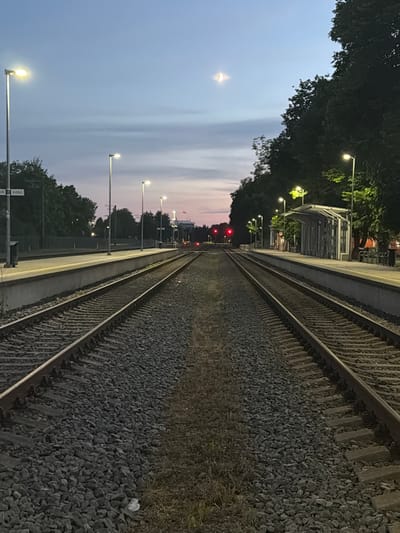 The image depicts a railway station at dusk, with two sets of train tracks stretching into the distance. Streetlights illuminate the platforms, and a few trees line the background. The sky is a soft gradient of blue and pink, with a crescent moon visible. The scene appears to be quiet and peaceful, with no people or trains present. The location is identified as the Sigulda train station in Sigulda, Latvia.