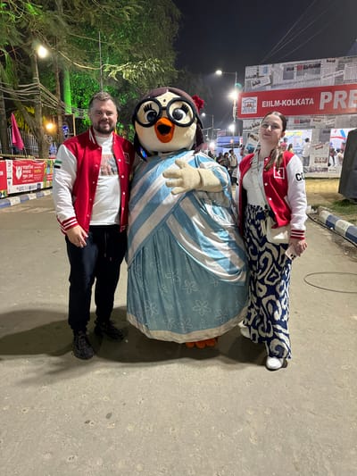 This image captures a nighttime scene at a public event in Bidhannagar, India, likely the International Kolkata Book Fair. Three individuals are positioned centrally, posing for a photograph.

On the left, an adult male with a beard and short dark hair wears a red and white varsity jacket over a white t-shirt, dark trousers, and dark athletic shoes. His left arm is around the central mascot.

The central figure is a costumed mascot resembling an anthropomorphic owl or penguin. It features large black-rimmed glasses, a red flower accessory on its head, a bindi, and an orange beak. The mascot is dressed in a light blue saree-style garment with white diagonal stripes on the bodice and white floral patterns on the skirt. Orange feet are visible at the base. Its right arm is slightly raised.

On the right, an adult female with dark hair wears a red and white varsity jacket over a white top, blue and white patterned wide-legged trousers, and white flat shoes. A white cross-body bag is slung over her shoulder, and she holds a light-colored smartphone in her left hand. Her right arm is around the central mascot.

All three subjects are looking directly at the camera, with the man and woman smiling. The ground is concrete. The background reveals an outdoor fair or exhibition setting illuminated by artificial lights. Trees with green lights are visible on the far left. In the middle-right background, a large banner displays "EIILM-KOLKATA" along with Bengali text that includes "KOLKATA BOOK FAIR". Crowds of people are discernible in the distance, and a blue and white painted curb runs along the right side of the pathway.