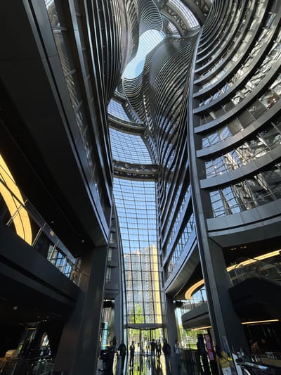 The image depicts an upward-angled view of an architectural structure in Fengtai District, China. The building features an interior with curved, layered tiers and prominent glass panels that allow natural light to filter in. The design incorporates linear elements and reflective surfaces. Reflected figures are visible on the right side of the image, suggesting a public space or walkway. The architectural style is modern and abstract, emphasizing the flow of light and the play of forms.