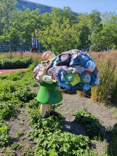 The image depicts a sculpture of a child carrying a large bundle of colorful, abstractly shaped objects, resembling discarded items or waste, on their back. The child figure is simplified, with a green dress and visible legs. The sculpture is situated outdoors in a landscaped area with gravel pathways and green vegetation. In the background, a modern building with multiple windows and trees with lush green foliage are visible. There is also a hint of a red track or path to the left of the sculpture. The overall impression is one of urban art in a park-like setting, potentially conveying a message about consumerism or environmental issues. This sculpture is located in the city of Rijswijk, Netherlands.