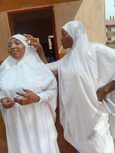 Two women in white robes and head coverings stand outdoors near a building. One woman smiles and holds a small pink and white item in her hands, while the other looks downward. The building is light tan or beige, and a window is visible to the left. Part of a building is also visible in the background. The image is rotated, giving a slightly off-kilter perspective.