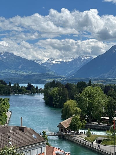 This is an aerial view captured on a sunny day with scattered clouds of the Aare River flowing through the city of Thun, Switzerland. The river, a striking turquoise color, is bordered by lush green trees and buildings with red-tiled roofs. In the foreground, a building with a tiled roof and a chimney is visible. A bridge crosses the river, with several people walking across it. Further in the distance, the turquoise river flows into a larger body of water, possibly Lake Thun, with sailboats dotting its surface. Majestic snow-capped mountains form the backdrop, adding to the scenic beauty of the location. The Aare River is a prominent natural feature of Thun, serving as a vital waterway and contributing to the city's picturesque landscape.
