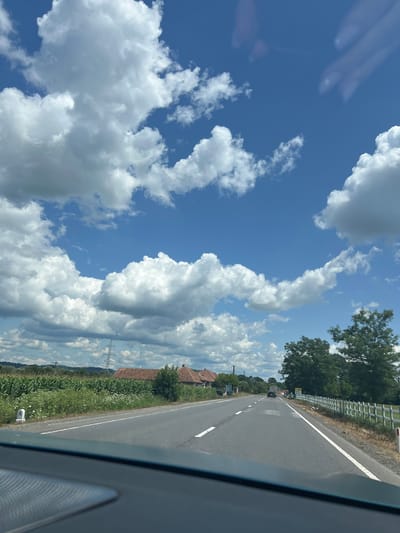 A paved, two-lane road stretches into the distance under a bright blue sky dotted with fluffy white cumulus clouds. The perspective is from inside a vehicle, looking forward. To the left of the road, a field of tall green corn is visible, interspersed with white wildflowers. Several red-roofed houses with brown tile are nestled behind the field, some with visible chimneys. A tall electricity pylon stands in the background. To the right of the road, a white picket fence runs along the edge, and a line of large green trees are prominent. A few cars are driving on the road ahead, and a truck is partially visible further down. The overall scene suggests a rural setting in Sâmbăta de Jos, Romania, on a sunny day.