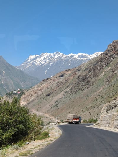 A truck, covered with a beige tarp, drives on a paved, winding road through a mountainous landscape. The truck is dark blue with an orange and red front. It is traveling on the right side of the road. The road curves from the foreground into the background, bordered by a steep, rocky mountainside on the right and a gentler, grassy slope on the left. In the distance, snow-capped mountains are visible under a clear blue sky. The location is the city of Gondhla, India.
