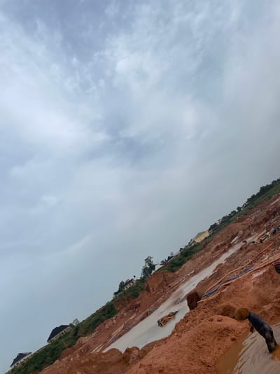 The image depicts a landslide in Rayfield, Nigeria, with a steep, muddy incline. Several houses are visible on the hillside above the landslide. The ground is covered in red soil and water, with some debris and construction materials scattered around. Orange and blue pipes are laid out across the terrain. There are several individuals present, some appearing to be working in the muddy conditions. One person is seen partially submerged in the mud. The sky is overcast with clouds.