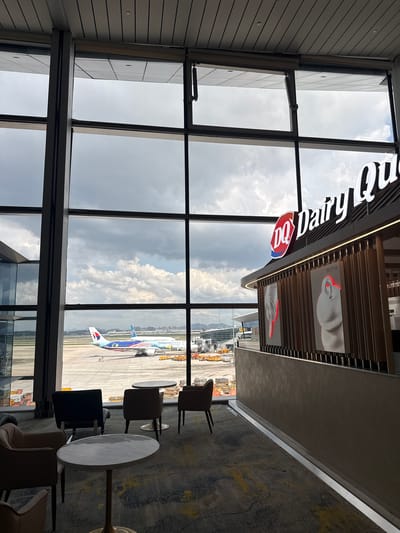 The image shows an indoor seating area at an airport with large windows overlooking the tarmac. A Dairy Queen restaurant is visible to the right, with its logo and decorative panels displaying images of ice cream. Outside the window, a Malaysia Airlines airplane is parked at a gate, with other aircraft and airport equipment in the background. Several chairs and tables are arranged in the seating area, suggesting a waiting lounge. The sky is cloudy, with a hint of mountains in the distance. The overall scene depicts a commercial airport environment. The location is identified as Baiyun District, China.