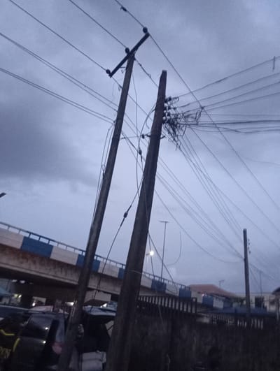 The image depicts a cloudy, overcast sky above a street scene in Port Harcourt, Nigeria. Two prominent wooden utility poles, laden with a dense network of electrical wires, dominate the foreground. The pole on the left is tilted, suggesting potential instability. To the left and behind the poles, a section of an elevated highway or bridge is visible, characterized by white and blue railings. Below the bridge, several dark-colored vehicles are parked or stopped, partially obscured by the utility poles and the low light conditions. The overall atmosphere is somber, possibly indicating twilight or an impending storm. The scene captures a common urban landscape in Port Harcourt, highlighting infrastructure and the dynamic interplay of man-made structures against the natural elements.