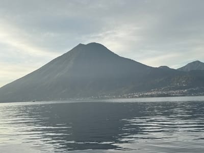 The scene depicts a panoramic landscape viewed from or near San Pablo La Laguna, Guatemala, looking across a wide body of water identified as Lake Atitlán. The lower half of the image is dominated by the lake's surface, which exhibits subtle ripples reflecting an overcast sky. In the distant midground, a small, dark vessel, possibly a boat or ferry, is visible on the water's surface towards the left.

The upper half of the image features a large, dark, conical mountain or volcano, likely Volcán San Pedro, rising prominently from the distant shoreline, its slopes appearing hazy and covered in dense, dark vegetation. To the right of this dominant peak, smaller mountains recede into the background, and a cluster of white and light-colored buildings, indicating a town or settlement, is visible along the base of these mountains at the distant water's edge. The sky above is light grey with diffuse clouds, suggesting an overcast condition or soft morning/late afternoon light. No individuals or explicit actions are discernible. The overall environment is serene and expansive.