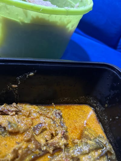 A close-up shot captured in Ilishan, Nigeria, displays two food containers on a blue fabric surface. In the foreground, a black rectangular plastic container holds a quantity of stew or soup. This dish features pieces of what appear to be meat or fish, immersed in a thick, reddish-orange, oily sauce. The sauce exhibits a slightly granular texture. Visible residue and droplets are present on the inner rim of the black container. In the upper background, partially obscured, is a light green, translucent bucket-like container. The top portion of this green container holds a light-colored, possibly granular or semi-solid substance, while the lower visible section contains a lighter-colored liquid. No individuals are present in the frame.