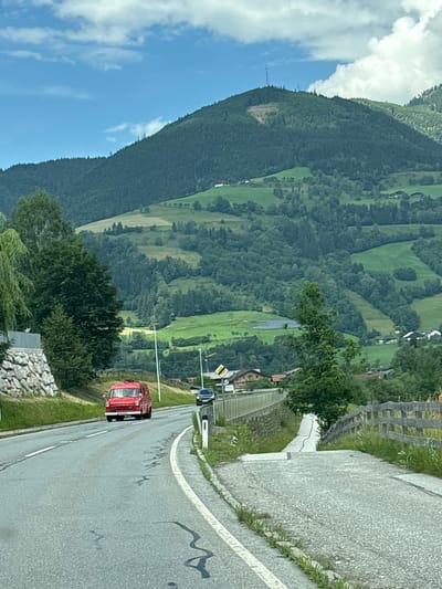 A red fire truck is driving on a paved road in a mountainous area. The road curves to the right, with a white line marking its edge. A wooden fence runs along the left side of the road, and grass and trees grow on either side. In the background, a lush green mountain slopes upward, covered in trees and dotted with fields and houses. The sky is blue with some white clouds. The scene appears to be set during the daytime, with fair weather. There are no people visible in the image. The location is suggested to be Bruck an der Großglocknerstraße, Austria, which is known for its scenic mountain landscapes.