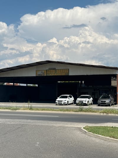 The image shows an outdoor scene featuring an auto repair shop. The building has a corrugated metal roof and open-sided bays where cars are parked. Three white cars are visible parked in a row under the covered area. A silver car is parked further back. A sign above one of the bays reads "AUTO SERVIS VUJAČIĆ". The setting appears to be a roadside location, with a paved road and a grassy verge running alongside the building. The sky is partly cloudy with bright sunlight, suggesting it is daytime, likely late morning or afternoon. The overall impression is that of a functioning business in a somewhat rural or suburban setting in Mojanovići, Montenegro.