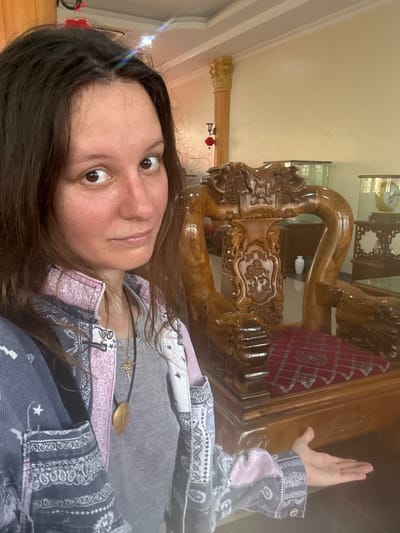 A medium shot captures a woman standing in what appears to be a museum or antique shop in Cương, Vietnam. She is positioned to the left of the frame, facing the camera with her head tilted slightly. The woman has long, dark brown hair and is wearing a grey t-shirt beneath an open, patterned jacket with shades of blue, white, and pink. A golden pendant hangs from a black cord around her neck. Her left arm is extended outwards, with her palm facing upwards, as if presenting something.
To her right, a large, intricately carved wooden chair dominates the center of the image. The chair is made of a reddish-brown wood, with detailed carvings of what appear to be dragons or mythical creatures. The seat of the chair is covered with a red cushion featuring a diamond pattern. Behind the chair and the woman, the background suggests an indoor environment with light-colored walls and wooden architectural elements, including a pillar to the left adorned with a decorative capital and a red lantern hanging from it. Various glass display cases and other furniture are visible in the background, reinforcing the idea of an exhibition space. The lighting is bright, suggesting daytime.