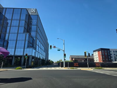 The image depicts a street view in Sacramento, California, USA. In the foreground, a paved road with white crosswalk markings is visible. A shadow of an object, possibly a vehicle or person, falls on the road from the left. To the left, a modern glass-fronted building, labeled "MAGGIE SQUARE," dominates the scene. A purple umbrella is partially visible on the far left. Traffic signals with green lights are prominent at the intersection, along with a "NO U-TURN" sign. Further into the scene, construction fencing with "WT WHITING-TURNER" branding is present. In the background, residential buildings with red and white facades are visible under a clear blue sky. The environment suggests an urban setting with ongoing development.