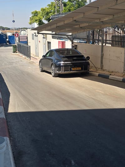 A black electric car, identified by its license plate as 713-90-603, is parked under a corrugated metal awning, connected to a charging cable. The car is a Hyundai Ioniq 6, distinguished by its streamlined design and distinctive rear light bar. The scene appears to be an outdoor charging station, likely in an industrial or service area, given the presence of large blue shipping containers and industrial fencing in the background. The environment is a paved area with a concrete curb marked with blue and white stripes. The ground is a mix of asphalt and dirt, with tire marks visible. The lighting suggests a sunny day, with strong shadows cast across the pavement. A red and white stop sign is visible on a pole near the entrance to the area. The location is identified as Beer Tuvia Regional Council, Israel.