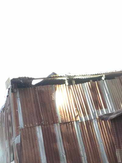 An upward shot captures a building's exterior, revealing corrugated metal cladding. Rust and tarnished silver stripes mark the horizontal panels. Sunlight streams through gaps, casting bright lines. The building's corner reveals edges with deteriorated metal and exposed structures. A bright white sky fills the background, creating high contrast.