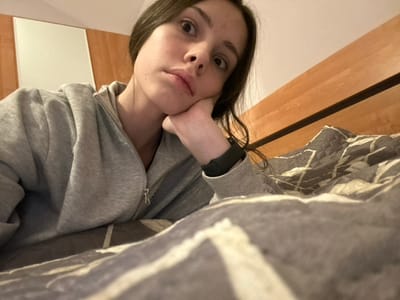 A person lies in bed, face close to the camera. She wears a grey zip-up hoodie. Her hand props her chin. Above, the bed's covers are visible, patterned with grey and white. The headboard is light-colored wood. The person's gaze is forward, expression neutral. A black smartwatch is visible on the wrist of the supporting hand. The photo is oriented in a disorienting way, as if the viewer is lying next to the subject.