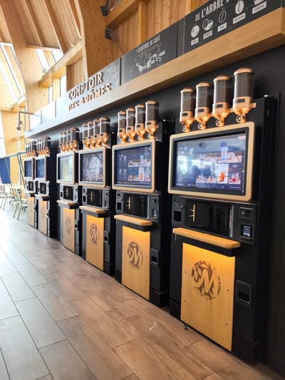 This media file captures a row of automated coffee vending machines in a modern, well-lit interior. The machines are predominantly black with warm wooden accents and feature digital touch screens displaying various coffee options. Above the screens, clear canisters hold coffee beans, ready for dispensing. Each machine is branded with a distinctive "M" logo surrounded by a coffee leaf motif, also etched into the wooden panels below the screens.

The wall behind the machines is dark, adorned with signage related to coffee production, including a map of the "Coffee Belt" and stages of coffee processing such as "Récolte," "Fermentation et Séchage," "Triage," "Torréfaction," and "Extraction." The ceiling is constructed of light-colored wooden beams and panels, giving the space an airy and natural feel, with large windows allowing ample natural light to flood the area.

The floor is covered in wood-look tiles. To the left of the vending machines, in the background, are tables and chairs, suggesting a waiting or seating area. The overall scene implies a self-service coffee station within a commercial establishment, possibly a cafe, a rest stop, or a food hall, emphasizing convenience and a modern approach to coffee consumption. There are no people visible in the frame. The setting appears clean and well-maintained.