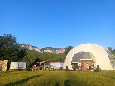 This image depicts an outdoor event scene at the "Dom Otdykha Zvezda" in Golubinskoye rural settlement, Crimea. The primary focus is a large, white geodesic dome structure serving as a stage. Inside the dome, three individuals are visible on a raised platform. One person is playing a guitar, while another appears to be operating equipment. A banner displaying a portrait is placed behind them. In front of the dome, two large black speakers are positioned on the grassy ground. To the left of the dome, a turquoise bus is parked, and a small ATV is situated in front of it. Behind the bus, a wooden fence encloses a small area. Further left, two large rectangular banners with images and QR codes are displayed. The landscape behind the event setup features a dense green forest leading up to a rocky mountain range under a clear blue sky. The foreground consists of a grassy field, with shadows indicating the presence of sunlight.