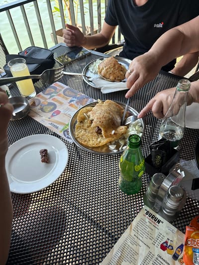 This is a close-up, eye-level outdoor shot of a table at a restaurant with a waterfront view, likely in Divic, Bosnia and Herzegovina. A group of people are engaged in eating and drinking. In the foreground, a white plate holds a single, small piece of dark brown meat. To the right, a silver metal salt and pepper shaker set sits on the table. A green glass bottle of "Limona" soda is positioned next to it. A black menu with "BURUN" and "Wifi. Restoran Burun" printed on it is also visible on the table. In the middle ground, a person's hands are visible holding a fork and knife, cutting into a dish of flatbread filled with meat and topped with chopped onions, served on a silver platter. Another silver platter with a similar dish is to the left, alongside a glass of light-yellow liquid and an ashtray. In the background, a person in a black t-shirt is seated, with their hands resting on the table. A black bag with a red zipper is placed on the table. The table surface is a black, perforated metal mesh. A metal railing and calm water are visible in the background, suggesting a waterside location.