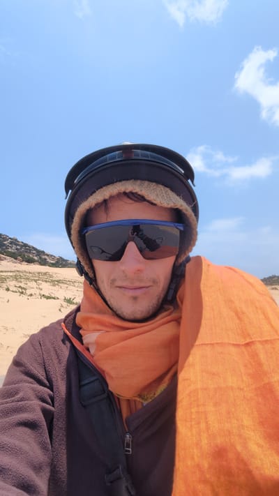 A male person is wearing sunglasses and a helmet, potentially participating in an outdoor activity such as paragliding. The background consists of a light blue sky with some clouds and a landscape with grassy terrain or dunes. The location is likely near Phuoc Dinh, Vietnam, based on the environment and the general appearance. The person is also wearing an orange scarf and layered clothing, including a brown fleece and a black strap.