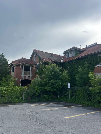 The image shows the exterior of an abandoned brick building covered in ivy, with a clay tile roof and a balcony supported by columns. The building is surrounded by lush green trees and vegetation, and a chain-link fence runs in front of it. A cracked asphalt parking lot with yellow parking lines is visible in the foreground. The sky is overcast and gray. A sign with text and a logo is posted on a pole near the fence. The scene appears to be located in Sainte-Anne-de-Bellevue, Canada.