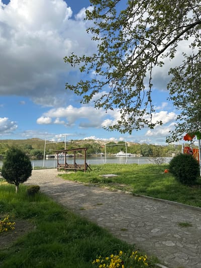 The image depicts a park setting in Beloslav, Bulgaria. A stone path curves from the lower right towards a white metal railing overlooking the water. A metal structure sits between the path and the railing. The scene includes green grass and bushes on the right and trees with green leaves framing the upper left. The sky is visible behind the trees, with blue patches and white clouds, with some dark, heavy clouds visible.