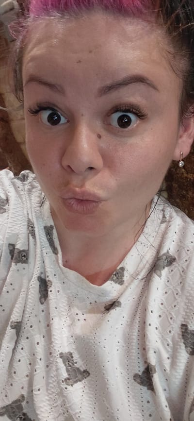 The image shows a close-up, head-and-shoulders shot of a woman with bright pink hair. She is looking directly at the camera with wide, surprised eyes and her lips are pursed in a "kissing" expression. She is wearing a white shirt with a detailed pattern of small gray teddy bears and eyelet cutouts. A small, sparkling earring is visible in her right ear. The background is out of focus, but appears to be a textured wall with brown and reddish tones. The woman has fair skin with some freckles and a mole on her cheek. The lighting is bright and appears to be from a frontal source, casting a slight shadow under her chin. The image was taken in Marynivka, Ukraine.