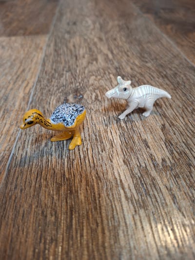 Two small, plastic dinosaur figurines are placed on a wooden surface with a wood grain pattern. The figurine on the left is yellow and orange with a speckled black and white back. It is depicted in a crouching position. The figurine on the right is a pale, off-white color with light blue stripes. It is standing on all fours and appears to be moving. The lighting in the image is soft, creating some shadow and highlights on the wooden surface. There are no people or other discernible objects in the scene. The setting does not provide any specific location information for Gorkovskiy, Russia.