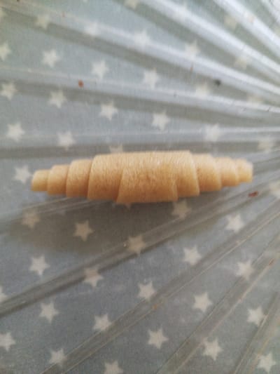 A close-up, overhead shot shows a single, tan-colored, spiral-shaped object lying horizontally across a pale blue surface patterned with white stars. The object appears to be made of a rolled, layered material, possibly a pastry or fruit leather, and tapers slightly towards its ends, resembling a miniature croissant or rolled fruit roll-up. The background surface has a matte finish and features an even distribution of five-pointed stars. The image is slightly out of focus and grainy, with some subtle variations in lighting and color. There is no discernible location in the city of Horsham, United Kingdom, visible in the image.