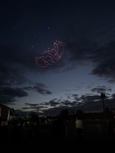 The image depicts a night scene in South Norfolk, United Kingdom. A firework display is visible in the dark sky, forming a pattern with pinkish-red bursts. The sky is also partially obscured by clouds. A tall structure, possibly a light tower, stands near the horizon. In the foreground, a crowd of people is present, with some individuals near a barrier or fence. Green lights illuminate part of the scene.