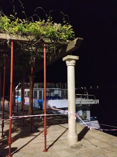 At night, the city of Peso da Régua, Portugal, features a waterfront scene. A partially visible, white and blue boat sits docked behind a low, open-air structure with a concrete column, which has red and white caution tape wrapped around it. Red, metal supports brace the wooden structure, which is overgrown with plants. The background is dark, indicating nighttime, with a few distant lights visible across the water. The stone pavement on the ground is illuminated by the ambient light.