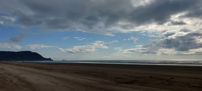 The image depicts a wide, sandy beach on the coast of Aleksandrovsk-Sakhalinskiy, Russia. The beach is mostly empty, with tire tracks visible in the sand, suggesting vehicle activity. In the distance, a dark, hilly landscape forms the coastline, with a prominent peak topped by a communication tower. The sea is calm, with gentle waves reaching the shore. The sky is a mix of blue and grey clouds, with sunlight breaking through in some areas, creating a bright reflection on the water. Three small, dark rock formations are visible protruding from the sea in the middle distance. The overall impression is a natural coastal scene under a partly cloudy sky.