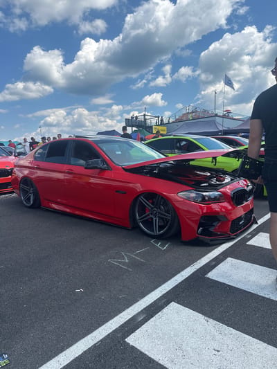 The image depicts an outdoor car show or event on a sunny day with a bright blue sky and scattered white clouds. The primary subject is a modified red BMW sedan with its hood open, showcasing the engine. The car is lowered and has custom dark gray wheels. In the background, several other cars are visible, including a bright green car with its hood also open. A crowd of spectators is gathered around the vehicles, some standing and talking, others observing the cars. Tents and structures, possibly vendor booths or event infrastructure, are present in the mid-ground. A flag with a blue emblem is flying on a pole behind the crowd. The ground is asphalt with white painted lines. The scene appears to be taking place in an open area, possibly a parking lot or event venue. The overall atmosphere suggests a casual gathering focused on automotive enthusiasm.