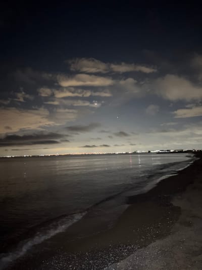 An aerial nighttime view of a coastal area in Chiba, Japan, captures the tranquil expanse of the ocean meeting the shore. The dark, choppy waters reflect the faint glow of distant city lights, creating a shimmering effect. The beach, composed of dark sand speckled with small shells, stretches into the foreground, with gentle waves lapping at the edge. The sky above is a deep, inky blue, dotted with wispy clouds illuminated by unseen light sources, and a couple of bright celestial bodies are visible, possibly stars or planets. The city lights stretch horizontally along the coastline in the middle distance, hinting at urban development beyond the natural landscape. The scene is serene and evokes a sense of peace and quiet.