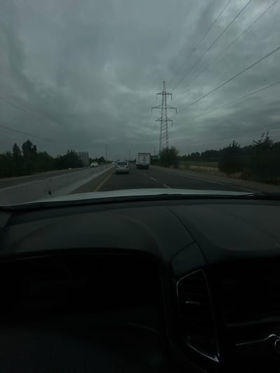 The image shows a view from inside a car, looking out at a highway under a cloudy sky. Several vehicles are on the road, including cars and a truck. There are power lines and a large transmission tower visible in the distance. Trees line the sides of the highway. The foreground of the image shows the dashboard and windshield of the car. The overall scene appears to be a typical highway landscape, possibly in a rural or semi-urban area, given the presence of trees and open space. The sky is overcast and gray, suggesting a potentially gloomy or rainy day. The light is diffused and shadows are not sharp. The scene is captured from a static point of view, implying the car is either stopped or moving slowly.