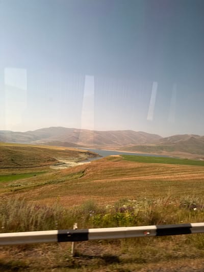 The image depicts a panoramic landscape scene, reportedly in Tsghuk, Armenia, viewed from a vehicle. The upper third of the frame shows a clear, light blue sky, transitioning to a hazy pale yellow at the horizon. Distant, undulating mountains with arid, muted brown and tan tones are visible along the horizon line. In the mid-ground, a calm, dark blue body of water, consistent with a reservoir or lake, curves from the left towards the right. Exposed, light-colored areas, indicative of dried lakebed or riverbed, are present along the water's edge on the left. Surrounding the water and extending into the foreground are diverse fields: a distinct vibrant green cultivated field lies to the right of the water, while the majority of the foreground and left mid-ground consists of dried, golden-brown grasses and fields. The immediate foreground is partially obscured by a white and black striped guardrail, running horizontally across the bottom fifth of the frame. Vertical light reflections are visible on the image, superimposed on the sky and distant mountains, consistent with a vehicle window. No human figures, animals, or explicit actions are present. The scene is illuminated by natural daylight, suggesting a dry period.