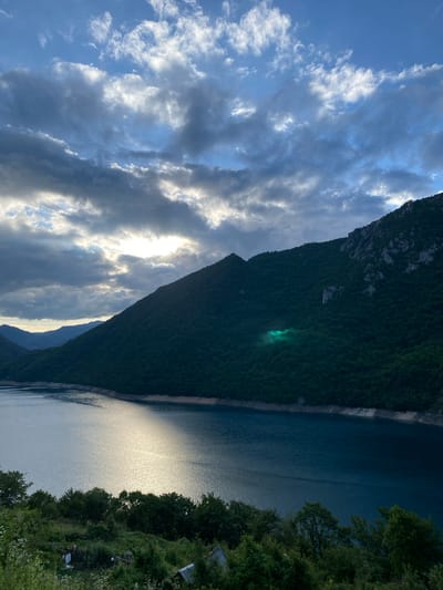 This expansive landscape photograph, taken in Plužine, Montenegro, captures a dramatic view of a large body of water, likely Lake Piva, nestled between steep, forested mountains under a partially cloudy sky.
The sky, occupying the upper left of the frame, is a blend of pale blue and dynamic, streaky clouds. Sunlight, breaking through the cloud cover, creates brilliant white highlights on the cloud edges and suggests either early morning or late afternoon.
A majestic mountain, densely covered in dark green trees and foliage, dominates the central part of the image. Its sheer face plunges into the water. A notable detail is a distinct, bright greenish glow emanating from a section of the mountain's forested slope, possibly a reflection or unique lighting.
The deep blue water, presumably Lake Piva, fills the lower right of the scene. Its surface beautifully reflects the low sun, forming a long, shimmering streak of light that cuts across the water's expanse. The far bank of the lake, on the mid-left, appears as a steep, lighter-toned natural wall.
On the immediate right, a thick fringe of green trees and bushes lines the water's edge. Peeking through this dense foliage are glimpses of small structures, suggesting houses or other buildings, indicating human presence along the lake's banks.
The overall scene evokes a sense of tranquil grandeur and natural immensity. No specific activities or individuals are visible. The lighting conditions point to either dawn or dusk, creating a soft yet powerful ambiance. There is no visible text in the image.