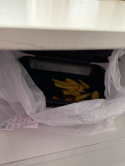 This media file depicts a close-up, overhead view of a white plastic bag containing food, specifically french fries, inside a black disposable container. The bag and container are placed within a white box or cooler. A crumpled receipt is partially visible, sticking out from the bag. The receipt shows Russian text and numbers, including "1.74 *1 шт. =231", "23 *1 шт. =82.2", "WT.=240.03", and "4.00". The overall scene suggests a takeaway food order. The lighting appears to be indoor, possibly natural light from above, and there are no indications of the time of day or weather. The location context is Uchaly, Russia.