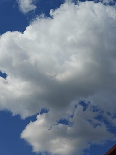 This is a vertical, medium-angle, outdoor photograph looking up at a predominantly blue sky filled with large, fluffy white and gray cumulus clouds. The clouds are textured and illuminated by sunlight, creating variations in brightness and shadow. The overall impression is of a partly cloudy day. The bottom right corner of the image shows a sliver of a dark red or brown roofline, suggesting the presence of a building. The image does not contain any people or visible evidence of specific human activity. The scene is likely located in Shkurupiivka, Ukraine, based on the provided context.