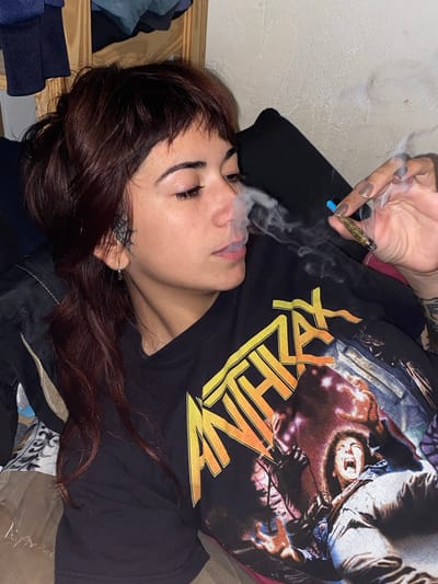 A person lies on their back wearing a black t-shirt with yellow and white graphics. Smoke is coming from their mouth as they hold a lit item in their hand. The person has dark hair and fair skin. The image was taken in an indoor setting with a wall in the background. The location is Monte Grande, Argentina.
