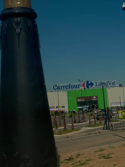 The image depicts an outdoor view of a Carrefour shopping center located in Beni Mellal, Morocco, under a clear blue sky. In the foreground, a dark, ornate pillar or lamppost with a golden finial occupies the left side of the frame, partially obscuring the midground. The ground level visible in the lower right consists of dry soil with sparse green vegetation.
In the midground, the Carrefour building features a white upper facade with prominent blue "Carrefour" and "LabelVie GROUPE" signage. The main entrance area is a vibrant green, displaying "مرحباً بكم" (Welcome in Arabic), "Bienvenue" (Welcome in French), and a smaller "Carrefour Centre Commercial Beni Mellal" sign. To the left of the green entrance, colorful advertisements are affixed to the white facade.
An access road or parking lot is visible in front of the building, featuring several rows of small palm trees planted in landscaped medians. Multiple vehicles, including cars and at least one motorcycle, are parked or in transit on the asphalt. A few individuals are discernible near the building's entrance and on the paved areas, indicating pedestrian activity. Dark streetlights are positioned throughout the parking area.
The background is dominated by a clear, unclouded light blue sky. A white dome-shaped security camera is mounted on a pole visible against the sky in the upper right quadrant of the image. A dark metal fence or gate structure is present in the lower right, separating the foreground from the shopping center's access.
