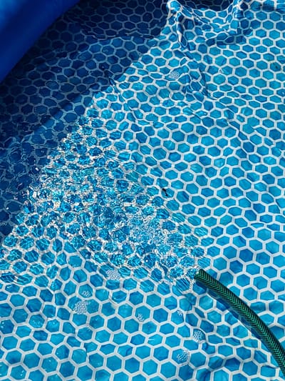The image shows the bottom of a kiddie pool with a blue and white hexagonal pattern. The pool is filled with water, and ripples are visible on the surface. A green garden hose is partially submerged in the water on the right side of the frame, suggesting the pool is being filled. The scene is brightly lit, likely by sunlight, creating shadows and highlights on the water and the pool liner. The location is specified as Mykilske, Ukraine, though the image itself does not provide any direct visual cues to confirm this specific location within the city.