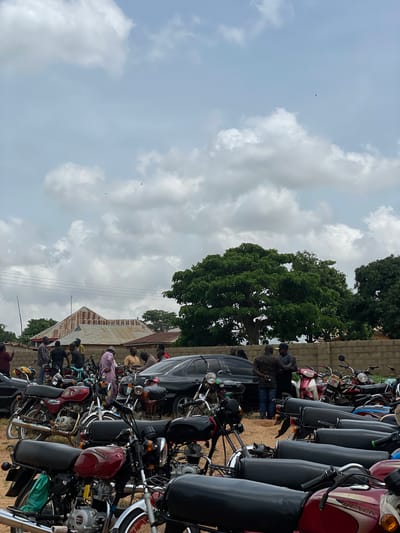 In Kassa, Nigeria, a gathering of people and parked motorcycles unfolds under a cloudy sky. In the background, a building with a rusty metal roof stands against the horizon, while lush green trees punctuate the landscape. The foreground is dominated by a collection of motorcycles, some red and black, parked closely together on a sandy surface. Several individuals are present, some standing near the motorcycles, others engaged in conversation. A black sedan is also visible, parked amongst the motorcycles. The scene suggests an informal outdoor meeting or event, possibly a community gathering or a market day.