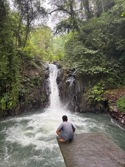 A person squats on a concrete platform extending over a fast-flowing river near a waterfall. Lush green trees and vegetation cover the surrounding rocky terrain. The water plunges over the rocks, creating a frothy, white cascade. The person, wearing a gray shirt, faces the waterfall, creating a contemplative scene amidst the natural environment. The image is rotated counterclockwise.